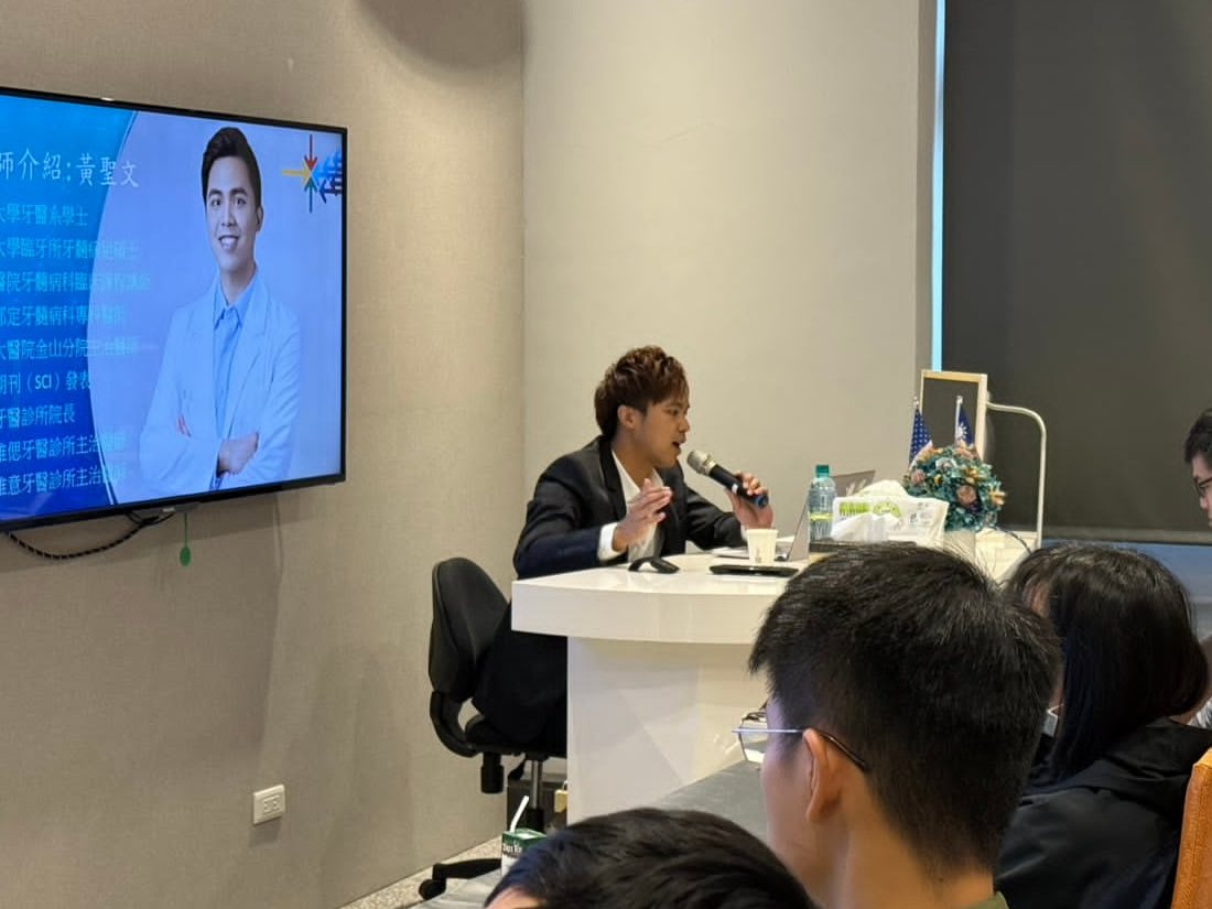 黃聖文醫師坐在講台前持麥克風授課，身後螢幕是講師介紹